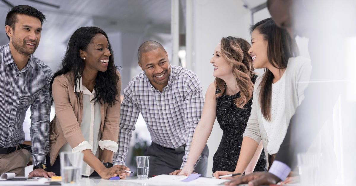 A multiethnic business team stands around the conference table to finalize a product launch. They are looking at each other and smiling or laughing as they wrap things up.