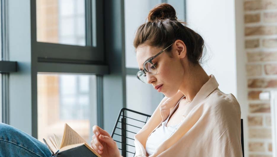 Image of focused beautiful woman in eyeglasses reading book while sitting in armchair at home